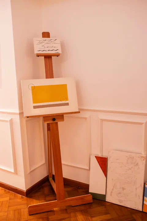 Cavalete de madeira com tela amarela e quadros apoiados na boiserie da galeria de arte
