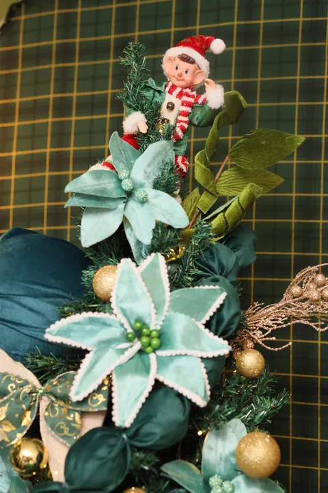 Elfo de Natal no topo de árvore com flores turquesa diante de parede xadrez verde
