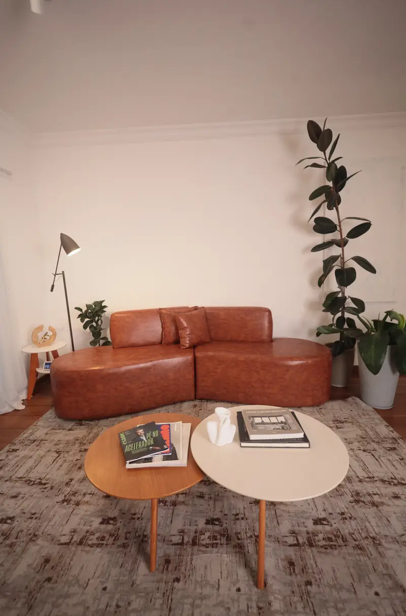 Sala do Loft Lumière com sofá curvo de couro caramelo, mesas de centro e figueira