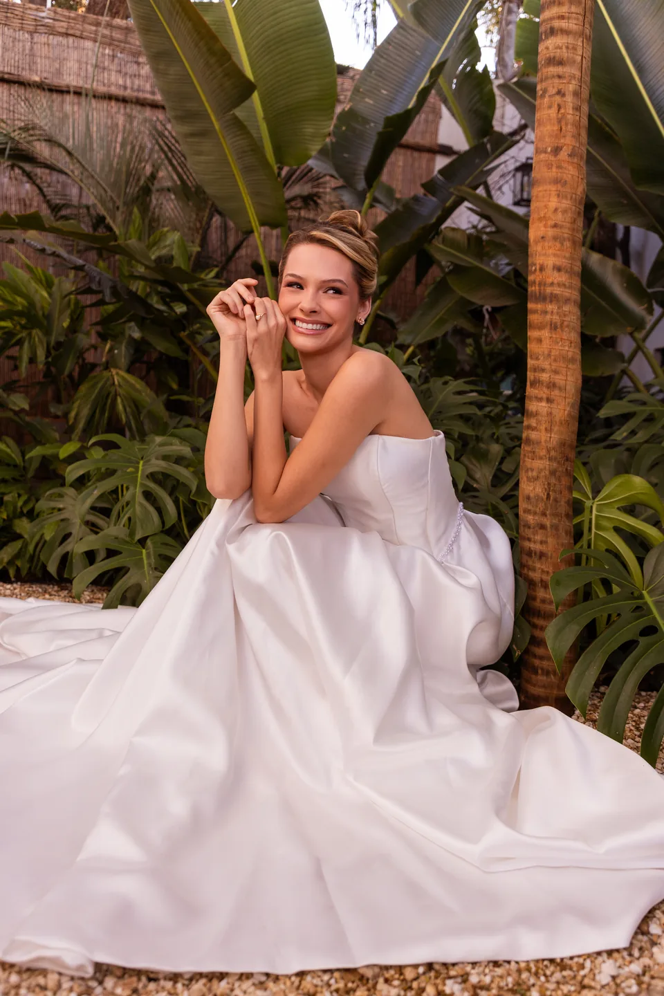 Noiva sentada no jardim, sorrindo, com o vestido espalhado sobre o cascalho