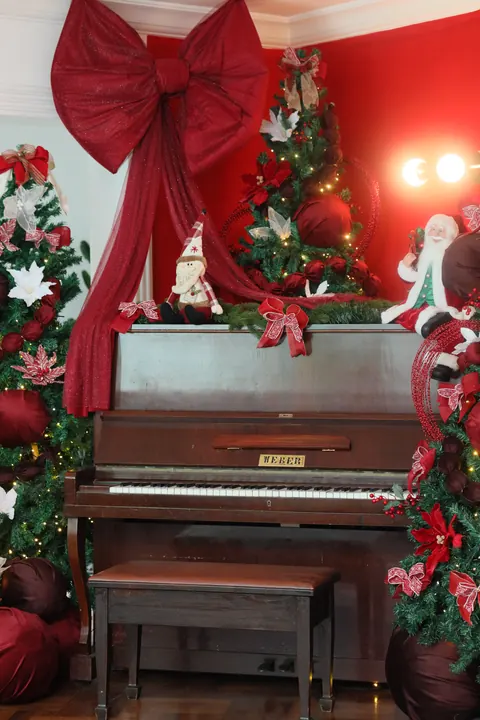 Piano antigo com grande laço vermelho e árvores de Natal no cenário vermelho do Loft Oásis